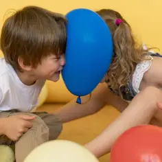 Dwoje dzieci z baloniem między twarzami, otoczeni kolorowymi balonami.
